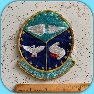 May include: A blue and turquoise embroidered patch with gold trim. The patch features a white eagle, a white book, and the text "Navy Student Squadron".