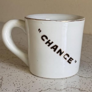 May include: A white ceramic mug featuring a curved handle and a metallic gold rim. The word "CHANCE" is printed in bold, dark, capitalized letters on the side of the mug, enclosed within quotation marks, positioned at a slight diagonal angle.