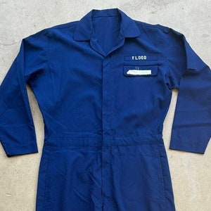 May include: A navy blue work jumpsuit with long sleeves and a zipper closure. The jumpsuit has a patch pocket on the chest with the word "FLOOD" printed on it.