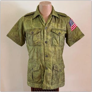 May include: A short-sleeved, quilted shirt in olive green camouflage pattern featuring a button-down front, two chest pockets, two lower pockets, and a small American flag patch on the left sleeve. The shirt has a collar and shoulder epaulets.
