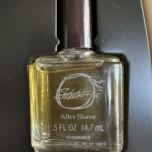 May include: A clear glass bottle of Stetson After Shave with a brown cap. The bottle contains 5 fl oz (14.7 ml) of liquid and is labeled as flammable. The bottle is sitting on a black surface.