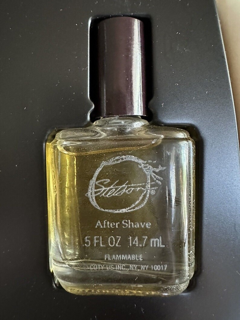 May include: A clear glass bottle of Stetson After Shave with a brown cap. The bottle contains 5 fl oz (14.7 ml) of liquid and is labeled as flammable. The bottle is sitting on a black surface.