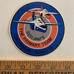 Puede incluir: Una pegatina redonda con un diseño azul, naranja y gris. Dos aviones blancos y rojos vuelan en formación. La pegatina dice "S211 JPATS The Primary Team Grumman - Augusta".