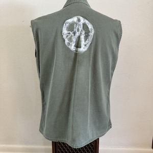 May include: Olive green sleeveless vest with a white peace sign painted on the back. The vest has a collared neckline and a button-down front. The peace sign is centered on the back and appears to be hand-painted.