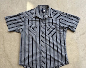 May include: A gray and black striped short-sleeved button-down shirt with a pointed collar and two chest pockets. The shirt has a Wrangler label.