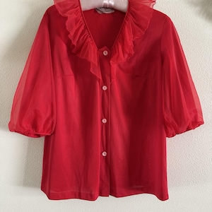 May include: A vibrant red blouse with a ruffled collar and sheer, three-quarter sleeves. The vintage top features a button-down front and is displayed on a hanger. The blouse is a solid color and appears to be made of a lightweight fabric.