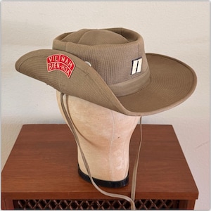 May include: A tan wide-brimmed hat with a chin strap. The hat features a red and white patch on the side with the text VIET-NAM BIEN-HOA and a small rectangular patch on the front with a black vertical bar on a white background.