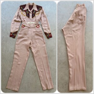 Pu&ograve; includere: Un completo pantalone vintage rosa chiaro con una camicia in stile western ricamata di marrone. La camicia ha ricami floreali sui polsini e sul collo. I pantaloni hanno vita alta e sono attillati.