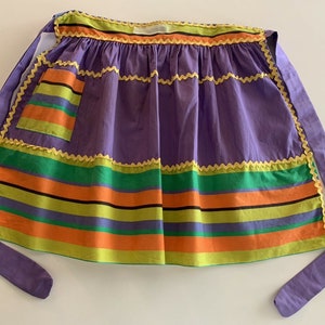 May include: A purple apron with a yellow, green, orange, and black striped bottom. The apron has a yellow scalloped trim and a single pocket.
