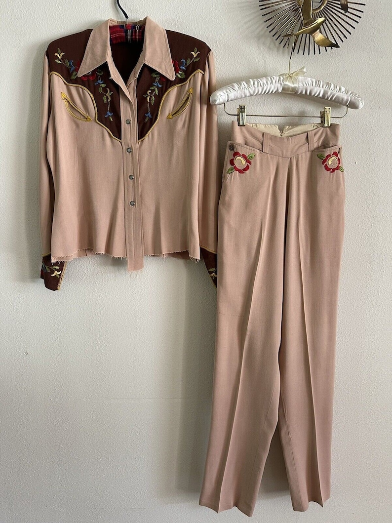 Pu&ograve; includere: Un completo in due pezzi di colore rosa chiaro. La parte superiore &egrave; una camicia a maniche lunghe con bottoni e ricami marroni sul collo e sui polsini. I pantaloni hanno vita alta e sono a gamba dritta con ricami floreali sulle tasche.