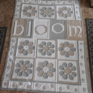 May include: A handmade, square baby blanket with a floral design. The blanket features the word "bloom" in tan letters. The blanket is white, tan, and gray, with a floral pattern in each square. The border has a floral and geometric design.