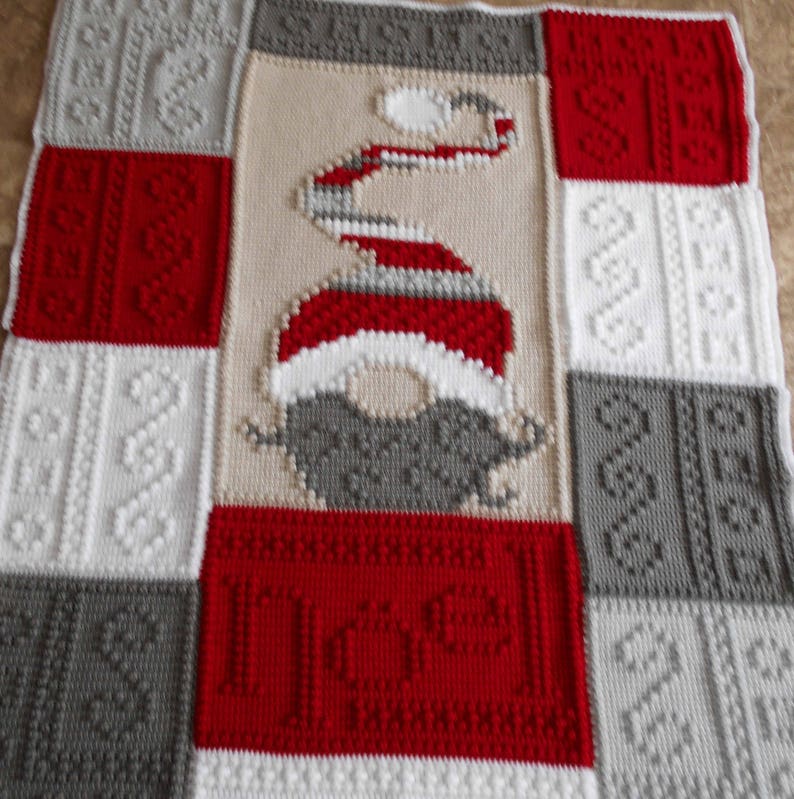 May include: A crocheted blanket with a red, white, and gray color scheme. The blanket features a gnome design in the center, with a red hat, white beard, and gray face. The blanket is made up of different squares with various textures.