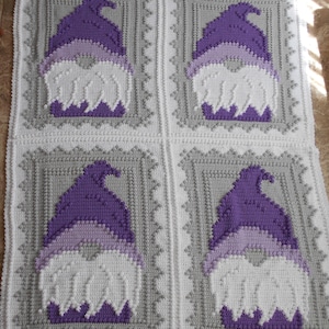 Peut inclure: Couverture crochetée avec quatre motifs de gnomes. Chaque gnome a un chapeau violet, une bande violet clair et une barbe blanche. La couverture est blanche avec des bordures grises et un bord décoratif.