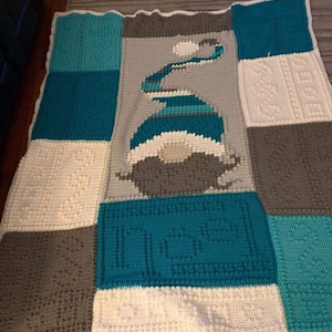 May include: A crocheted baby blanket with a blue and gray color scheme. The blanket is made up of squares with different patterns. The center square features a blue and white design of a snowman. The blanket is signed "Norma Ciotti Weiland".