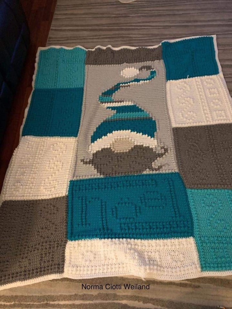 May include: A crocheted baby blanket with a blue and gray color scheme. The blanket is made up of squares with different patterns. The center square features a blue and white design of a snowman. The blanket is signed "Norma Ciotti Weiland".