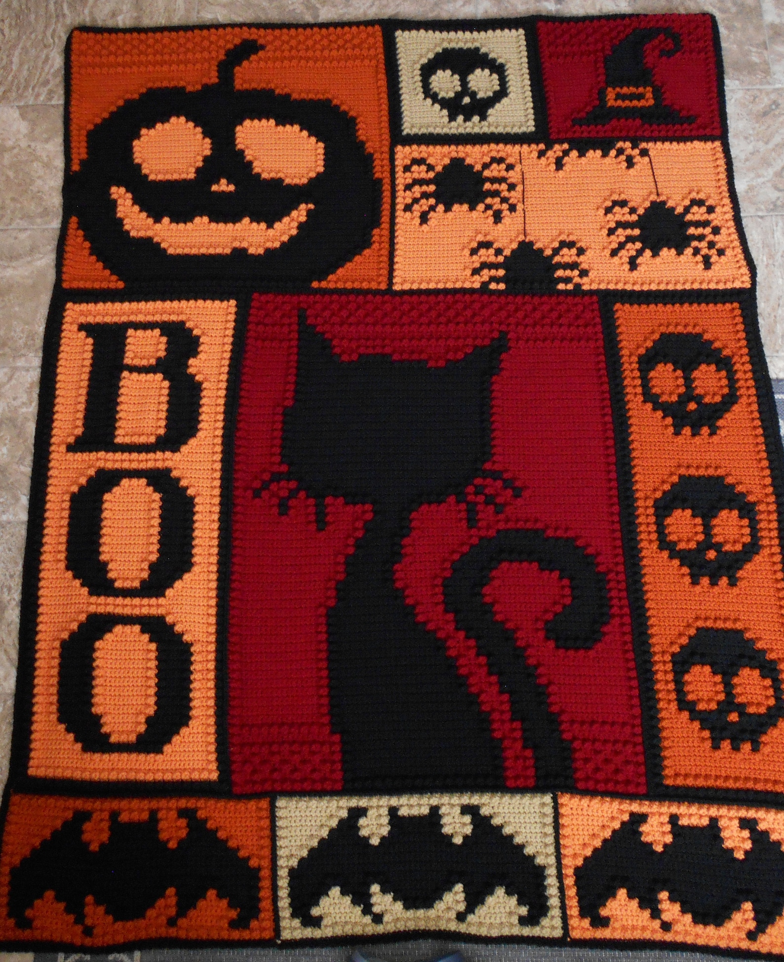 Crochet Halloween blanket with black cat, jack-o'-lantern, BOO letters, and spooky icons in orange, red, and black
