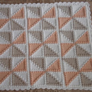 Puede incluir: Una manta de ganchillo hecha a mano con un patrón geométrico de triángulos en blanco, melocotón y topo. La manta tiene un borde festoneado y está hecha con un punto texturizado. Ideal para un bebé o como manta decorativa, mide aprox. 100 cm x 100 cm.