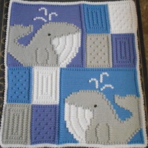 Può includere: Una copertina per neonati all'uncinetto con uno schema di colori blu, bianco e grigio. La copertina presenta due disegni di balene con getti d'acqua bianchi.