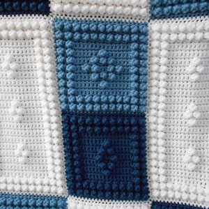 May include: A white crocheted blanket with a blue and white checkered pattern. The blanket is made with a textured stitch that creates a raised, bumpy surface.