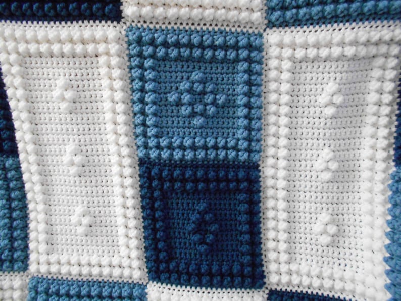 May include: A white crocheted blanket with a blue and white checkered pattern. The blanket is made with a textured stitch that creates a raised, bumpy surface.