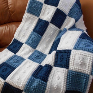 May include: A crocheted baby blanket with a checkered pattern in shades of blue and white. The blanket is made with a textured stitch that creates a raised, bumpy surface.