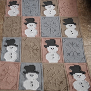 Puede incluir: Una manta de ganchillo con un patrón repetido de muñecos de nieve y copos de nieve. La manta está hecha en tonos de marrón, gris y rosa.