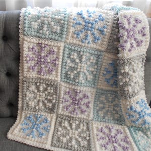 May include: A handmade crochet blanket featuring a snowflake design. The blanket is composed of square blocks in shades of white, gray, light blue, and lavender. The snowflakes are in contrasting colors, adding visual interest. The blanket is draped over a gray sofa.