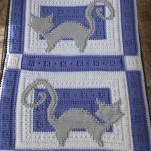 May include: A crocheted baby blanket with a white background and blue and gray stripes. The blanket features two gray cats with curled tails in the center.