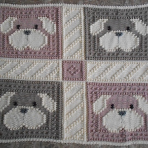 May include: A crocheted baby blanket with a repeating pattern of puppy faces in shades of pink, brown, and white.