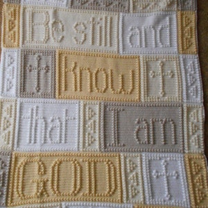 May include: A white crocheted blanket with the words "Be still and know that I am good" in beige and yellow yarn.