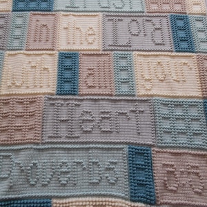 May include: A crocheted baby blanket with a blue, beige, and white color scheme. The blanket features the words "Trust in the Lord" in a raised stitch pattern.