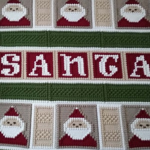 May include: A crocheted blanket with a green, white, and beige pattern. The blanket features a repeating pattern of Santa Claus faces and the word "SANTA" in white letters.