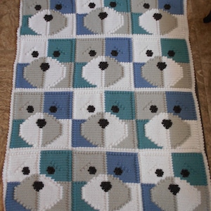 Può includere: Una copertina per neonati all'uncinetto con un motivo ripetitivo di quadrati bianchi, grigi, blu e turchese che presentano un viso di cane stilizzato in ogni quadrato.