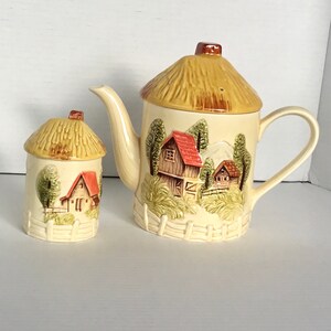 May include: A ceramic teapot and sugar bowl set with a cottagecore theme. The teapot and sugar bowl are decorated with a painted scene of a small village with houses, trees, and a fence. The lids of the teapot and sugar bowl are shaped like thatched roofs.