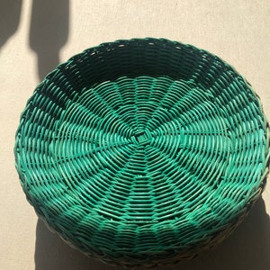 May include: A round, green wicker basket with a woven pattern. The basket is made of natural materials and has a rustic look.