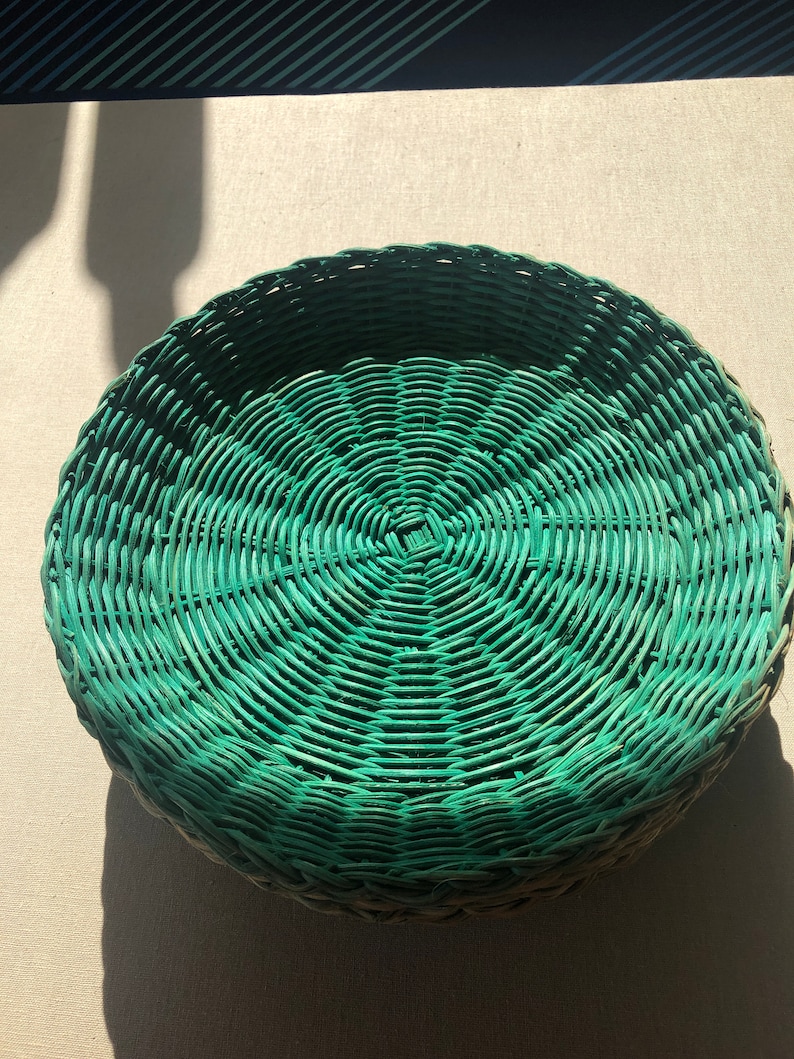 May include: A round, green wicker basket with a woven pattern. The basket is made of natural materials and has a rustic look.