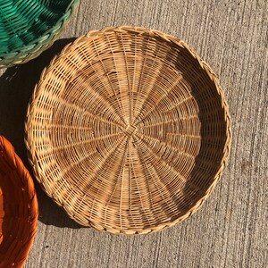 May include: A round, woven wicker basket with a natural brown color. The basket is made of thin, tightly woven strips of wicker, creating a smooth, even surface.