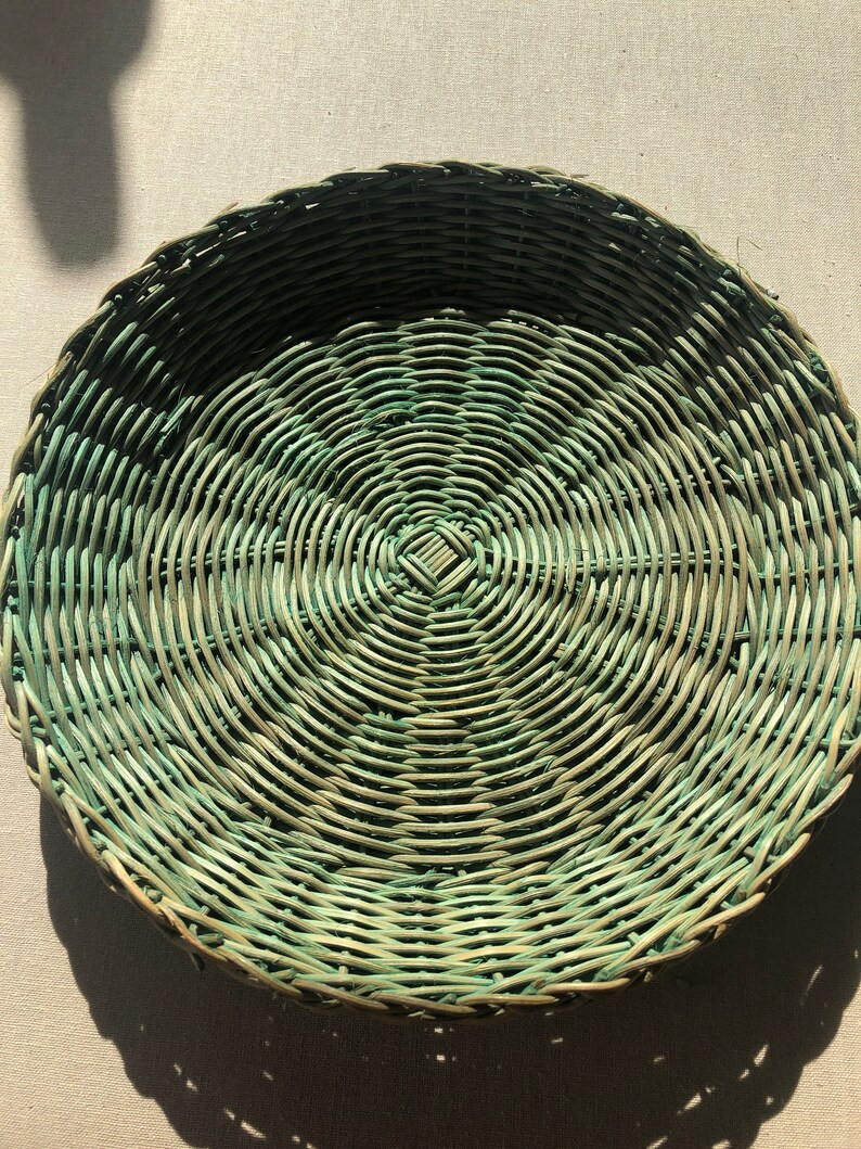 May include: A round woven wicker basket with a green and natural color pattern. The basket is made of natural materials and has a circular shape.