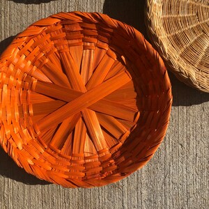 May include: A round, woven basket with a bright orange color. The basket is made of natural materials and has a unique, intricate design.