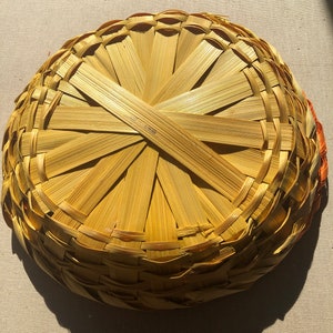 May include: A woven basket made of natural materials, with a circular shape and a light brown color. The basket is made of thin strips of woven material, creating a pattern of concentric circles.