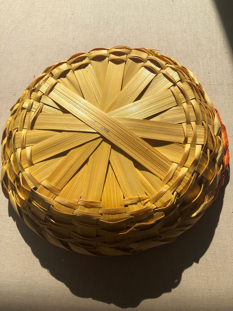May include: A woven basket made of natural materials, with a circular shape and a light brown color. The basket is made of thin strips of woven material, creating a pattern of concentric circles.