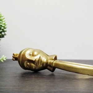 May include: A gold-coloured nutcracker shaped like a human face. The nutcracker has a long handle and a hinged jaw.