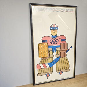 May include: Framed vintage poster for the Winter Olympics in Denver. The artwork features a stylized hockey goalie in red, blue, and tan, with the Olympic rings and the number 76. The goalie's helmet and stick have stars and stripes.