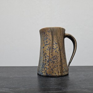 May include: A brown ceramic mug with a speckled glaze and a blue and brown color scheme. The mug has a rounded body and a handle.