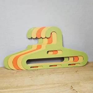 May include: Five brightly coloured plastic clothes hangers in a stack. The hangers are in shades of green, yellow, and orange. Each hanger has a distinctive design with a curved top and a rectangular body with cutouts.