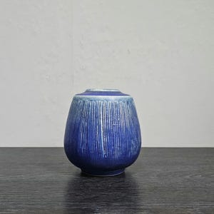 Könnte beinhalten: Eine kleine, blaue Keramikvase mit einer strukturierten, gerippten Oberfläche. Die Vase hat eine abgerundete, bauchige Form und eine leicht ausgestellte Öffnung.