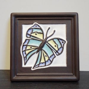 Könnte beinhalten: Eine Keramikfliese mit braunem Rahmen zeigt einen bunten Schmetterling mit blauen, gelben und grünen Flügeln. Der Schmetterling ist auf einem weißen Hintergrund zentriert.