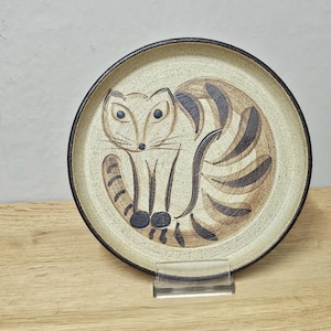 May include: A ceramic plate with a hand-drawn cat design. The cat is depicted in brown and black stripes, with large eyes and a long tail. The plate has a beige background and a dark brown rim.
