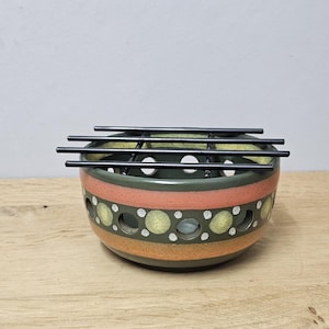 May include: A decorative ceramic bowl with a black metal grill. The bowl has a green interior and is decorated with orange, yellow, and white circular patterns. The bowl sits on a wooden surface.