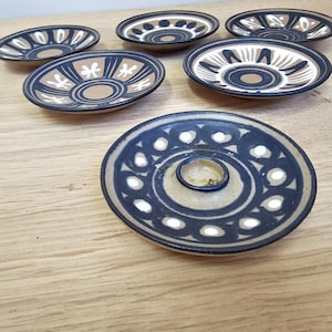 May include: A collection of ceramic dishes with a beige base and dark blue and white geometric patterns. The dishes vary in size and design, with some featuring a central raised area. The arrangement is on a light wood surface.